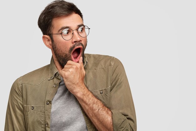 Scared anxious male keeps hand near opened mouth, looks with very frightened expression aside, notices something awful, wears round spectacles and fashionable shirt, poses in against white wall