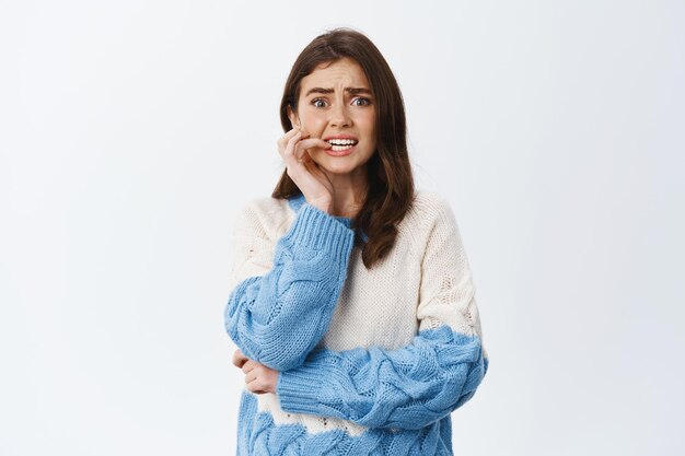 Scared and anxious woman standing worried and nervous, biting finger nail and frowning, trembling terrified, standing in sweater on white