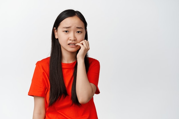Scared asian girl biting fingers and looking on white insecure, anxious about something, feeling worried, standing on white.