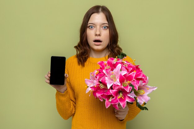 Scared beautiful young girl on happy woman's day holding bouquet with phone isolated on olive green wall