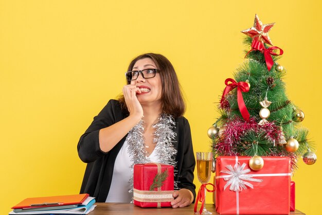 Scared business lady in suit with glasses holding her gift and sitting at a table with a xsmas tree on it in the office