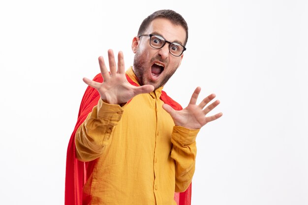 Scared caucasian superhero man in optical glasses with red cloak stands with raised hands isolated on white background with copy space