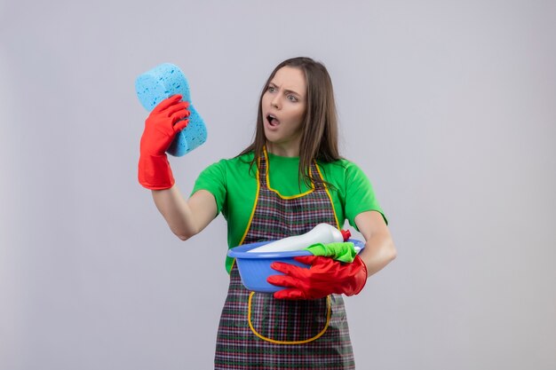 Scared cleaning young girl wearing uniform in red gloves holding cleaning tools looking at sponge on her hand on isolated white background