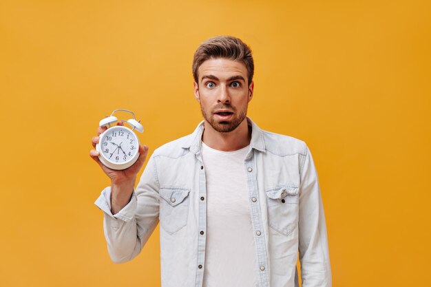 Scared cool guy with ginger beard and brown hair in stylish light outfit holding big alarm clock on isolated background