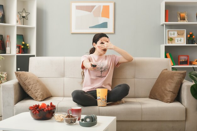 Scared covered eye with hand young girl holding tv remote, sitting on sofa behind coffee table in living room