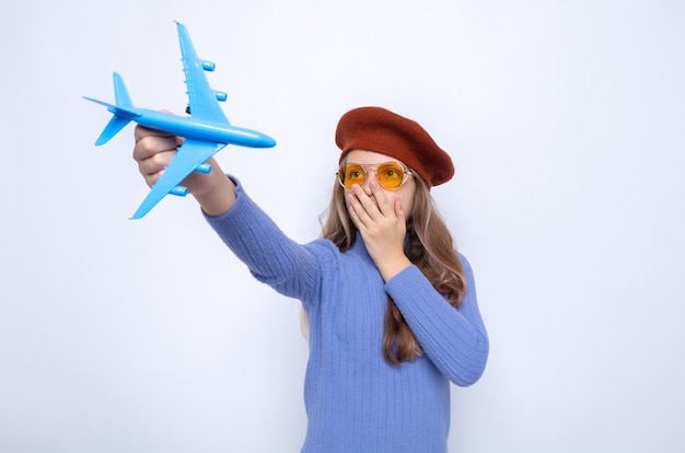 白い壁に隔離されたおもちゃの飛行機を差し出す帽子と眼鏡をかけている手で怖い覆われた顔美しい少女