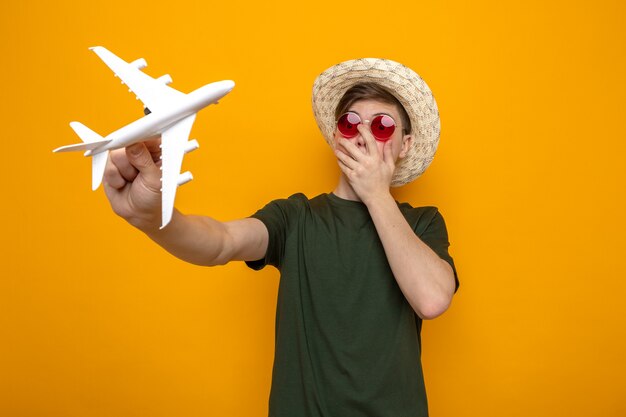 Испуганное закрытое лицо рукой молодой красивый парень в шляпе с очками, держащий игрушечный самолетик