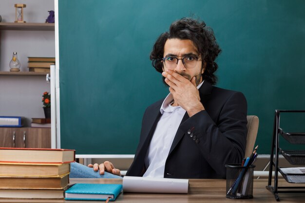 Scared covered mouth with hand male teacher wearing glasses sitting at table with school tools in classroom