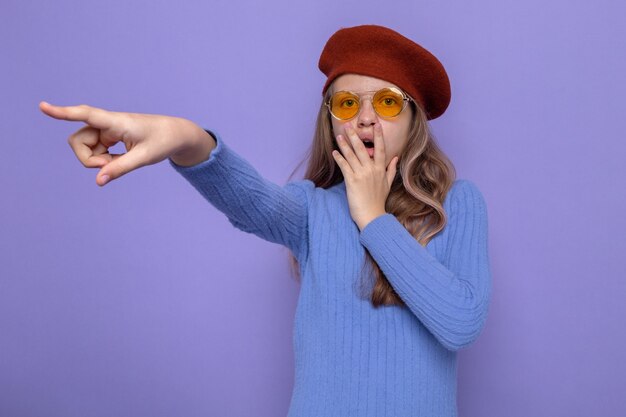 Scared covered mouth with hand points at side beautiful little girl wearing hat with glasses 