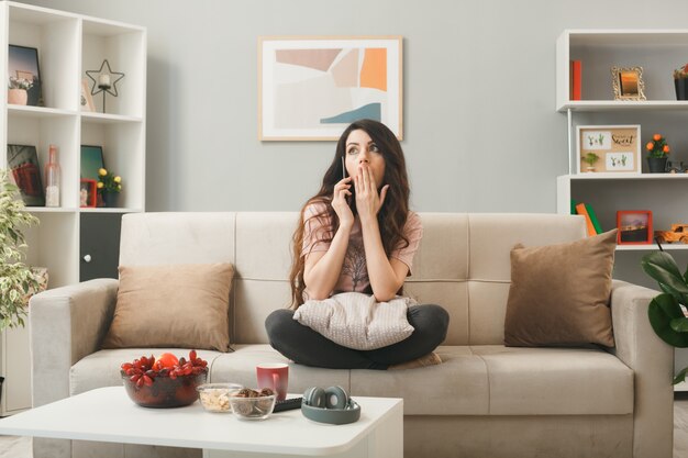 Scared covered mouth with hand young girl speaks on phone sitting on sofa behind coffee table in living room