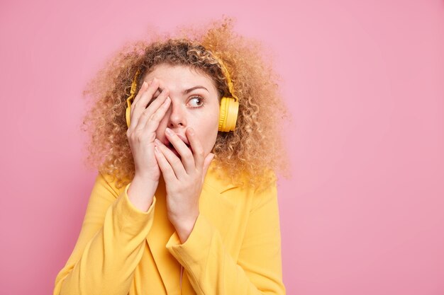 Scared curly haired woman feels very frightened covers face looks away 