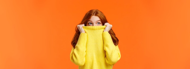 Scared cute teenage redhead girl pulling sweater on face and peeking at upper left corner with frigh