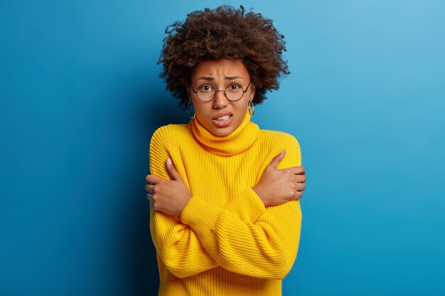 Scared dark skinned woman keeps hands crossed, trembles from fear, wears round spectacles and yellow sweater, isolated over blue background