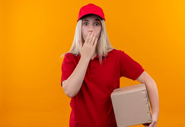赤いTシャツとキャップ保持ボックスを身に着けている怖い配達少女は孤立したオレンジ色の背景に彼女の手を口に置きます