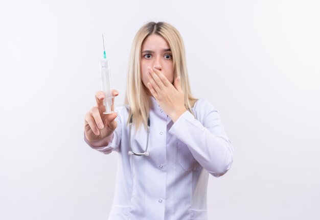 Scared doctor young blonde girl wearing stethoscope and medical gown holding syringe covered mouth on isolated white background