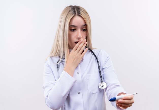 聴診器と医療用ガウンを身に着けている怖い医者若いブロンドの女の子は、孤立した白い背景の上の手で覆われた口の温度計を見て