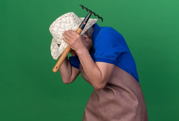 Free photo scared elderly female gardener wearing gardening hat puts hands on head holding rake on green