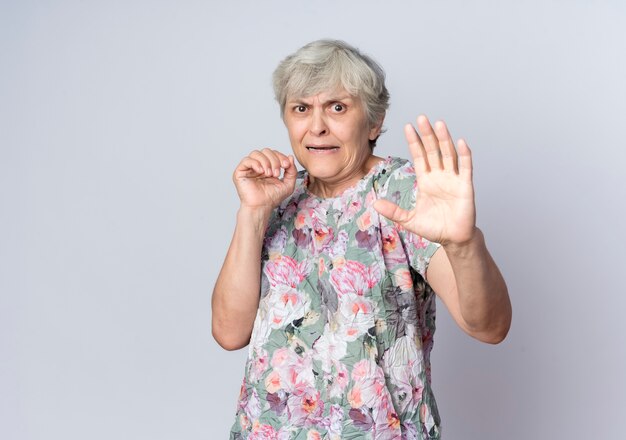 Scared elderly woman stands with raised hands isolated on white wall
