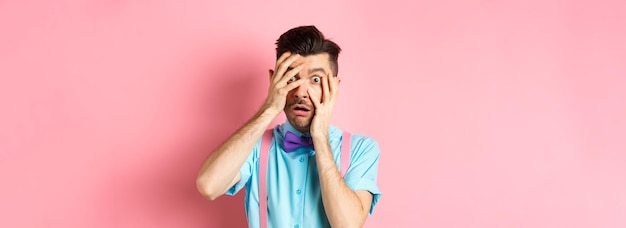 Free photo scared and embarrassed nerdy guy in bowtie covering his eyes peeking through fingers at something sc