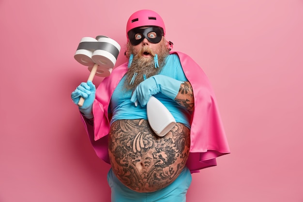 Испуганный эмоциональный бородатый мужчина-супергерой, ошеломленный тем, что выполняет слишком много задач, носит маску, шлем и плащ, резиновые защитные перчатки позирует с инструментами для чистки на розовой стене