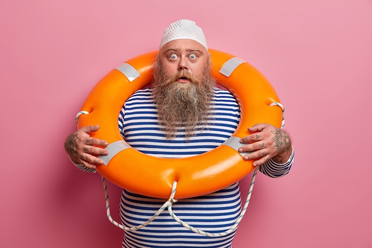 Free Photo scared emotional bearded man stares with shocked gaze, poses with lifering, wears striped sailor shirt, enjoys water recreation and summer holidays at seaside, isolated on pink wall