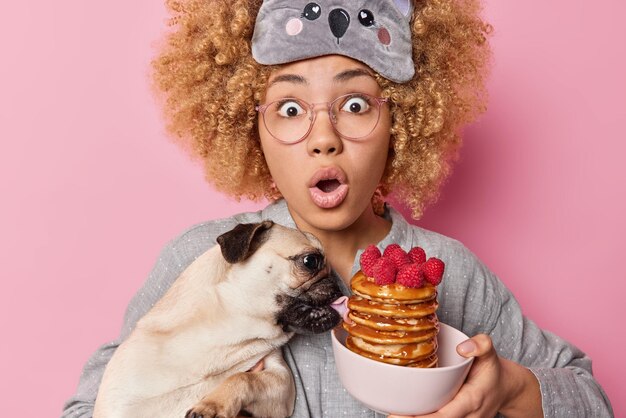 ピンクの壁に隔離されたナイトウェアに身を包んだ朝食用に作られたパンケーキをなめる衝撃ホールドパグ犬から口を大きく開いた巻き毛の凝視バグの目を持つ怖い感情的な女性