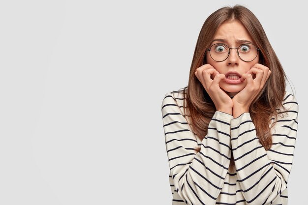 Scared European young female looks with bugged eyes, keeps hands on cheeks, dressed in striped sweater, isolated over white wall