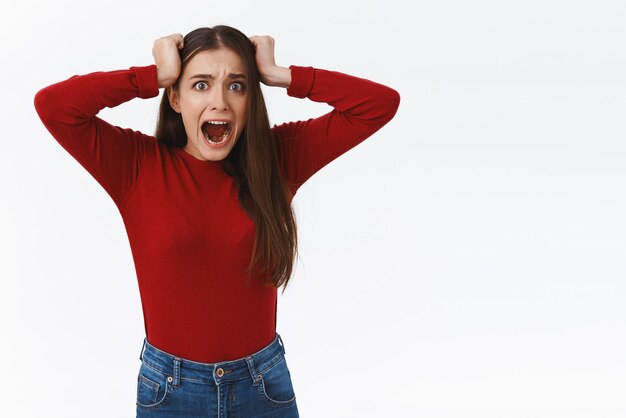 Scared freakedout panicking brunette girl in red sweater screaming from panic attack and fear pulling hair from head stare camera insecure standing troubled and horrified white background