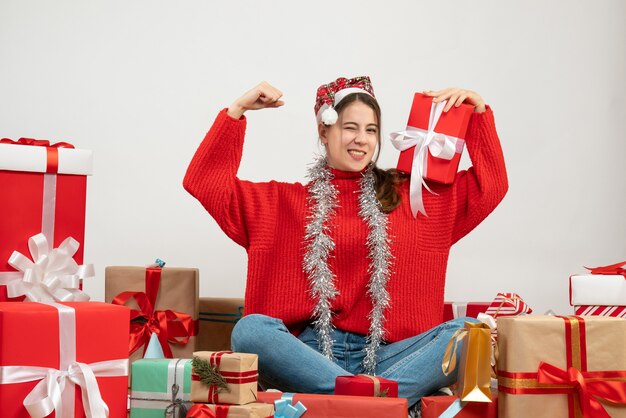 scared girl with santa hat showing winning gesture sitting around presents on white