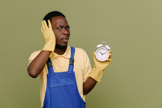 Scared grabbed head holding alarm clock young africanamerican cleaner male in uniform with gloves isolated on green background