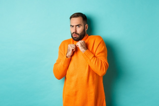 Scared guy jumping startled, looking at something scary, standing in orange sweater, over turquoise wall.