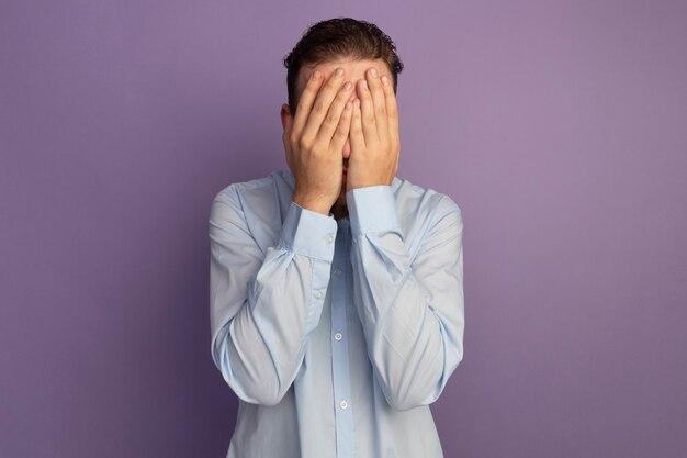 Scared handsome blonde man puts hands on face isolated on purple wall