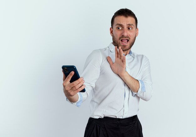 Scared handsome man holds and looks at phone with raised hand isolated on white wall