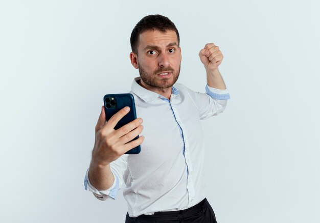 Scared handsome man holds phone and raises fist ready to punch isolated on white wall