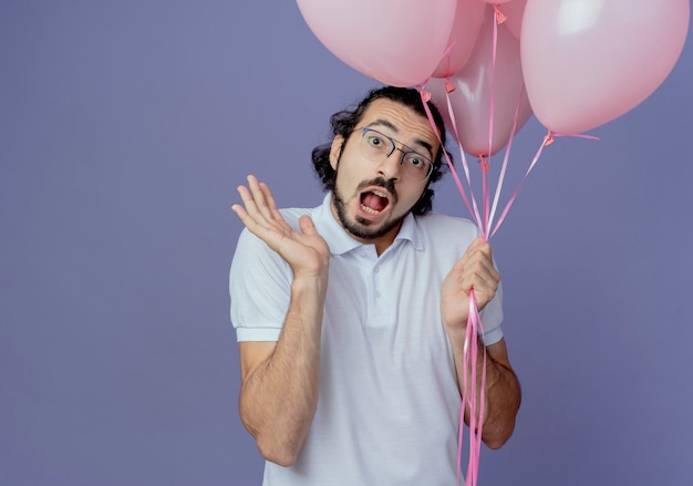 Free photo scared handsome man wearing glasses holding balloons and spread hand isolated on purple