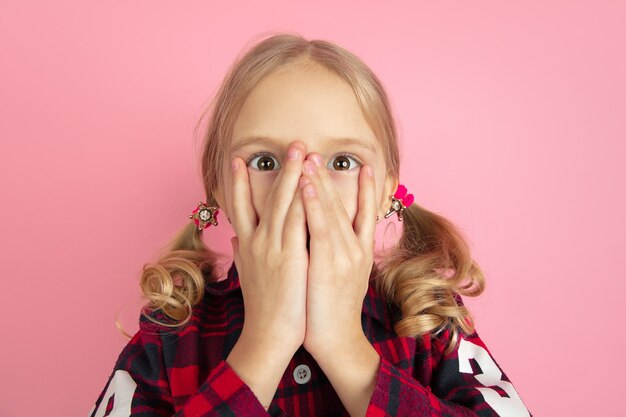 Scared hiding face. Caucasian little girl's portrait on pink wall.