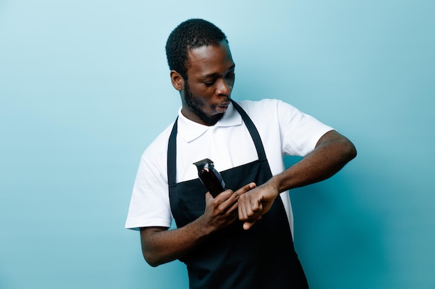 Scared holding hair clippers young african american barber in uniform isolated on blue background