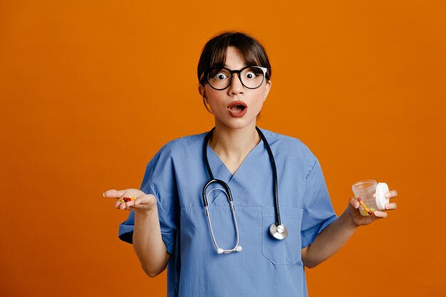 Spaventata tenendo la pillola e il contenitore della pillola giovane dottoressa che indossa uno stetoscopio fith uniforme isolato su sfondo arancione