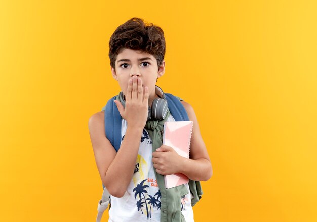 Испуганный маленький школьник в сумке на спине и наушниках держит ноутбук и прикрывает рот рукой, изолированной на желтом