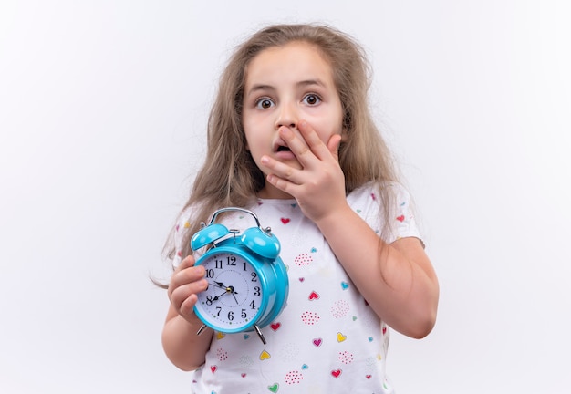 Scared little school girl wearing white t-shirt holding alarm clock covered mouth on isolated white background