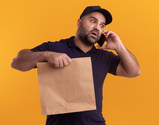 Scared looking at side middle-aged delivery man in uniform and cap holding paper food package speaks on phone isolated on yellow wall