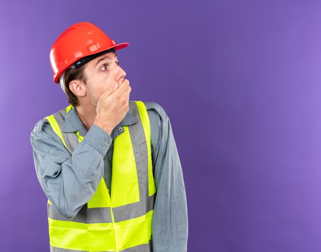 Scared looking side young builder man in uniform covered mouth with hand 