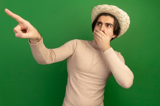 Free photo scared looking at side young handsome guy wearing hat covered mouth with hand points at side isolated on green wall