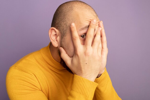 Scared looking straight ahead young handsome guy covered face with hands isolated on purple