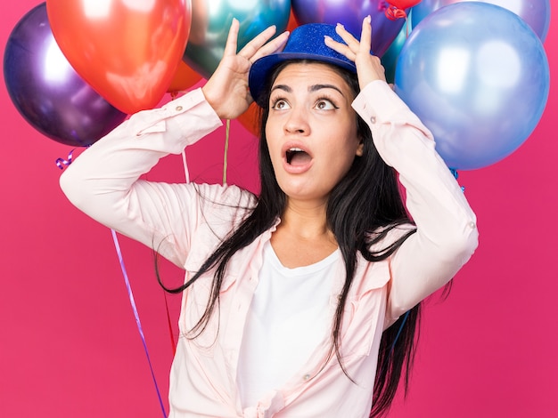 Scared looking up young beautiful girl wearing party hat standing in front balloons 