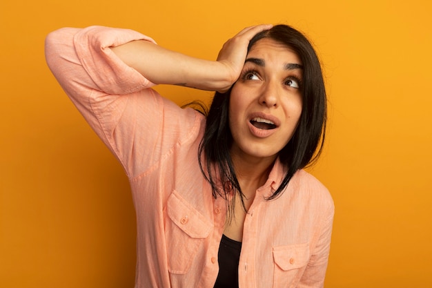 Scared looking up young beautiful girl wearing pink t-shirt putting hand on head isolated on yellow wall