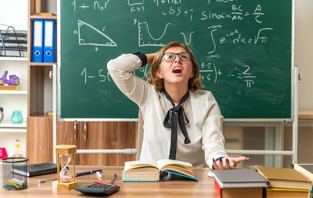 Free photo scared looking up young female teacher wearing glasses sits at table with school tools putting hand on head in classroom