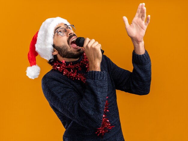 Scared looking up young handsome guy wearing christmas hat with garland on neck speaks on microphone points at up isolated on orange background