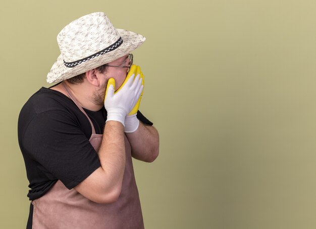 Scared looking at behind young male gardener wearing gardening hat and gloves covered face with hands isolated on olive green wall with copy space