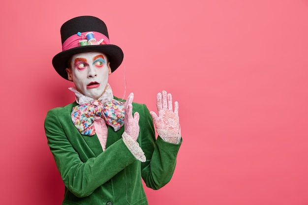 Free photo scared male hatter makes defense gesture afraids of something falling from height wears big hat lace gloves and green velvet jacket poses against rosy wall with blank space